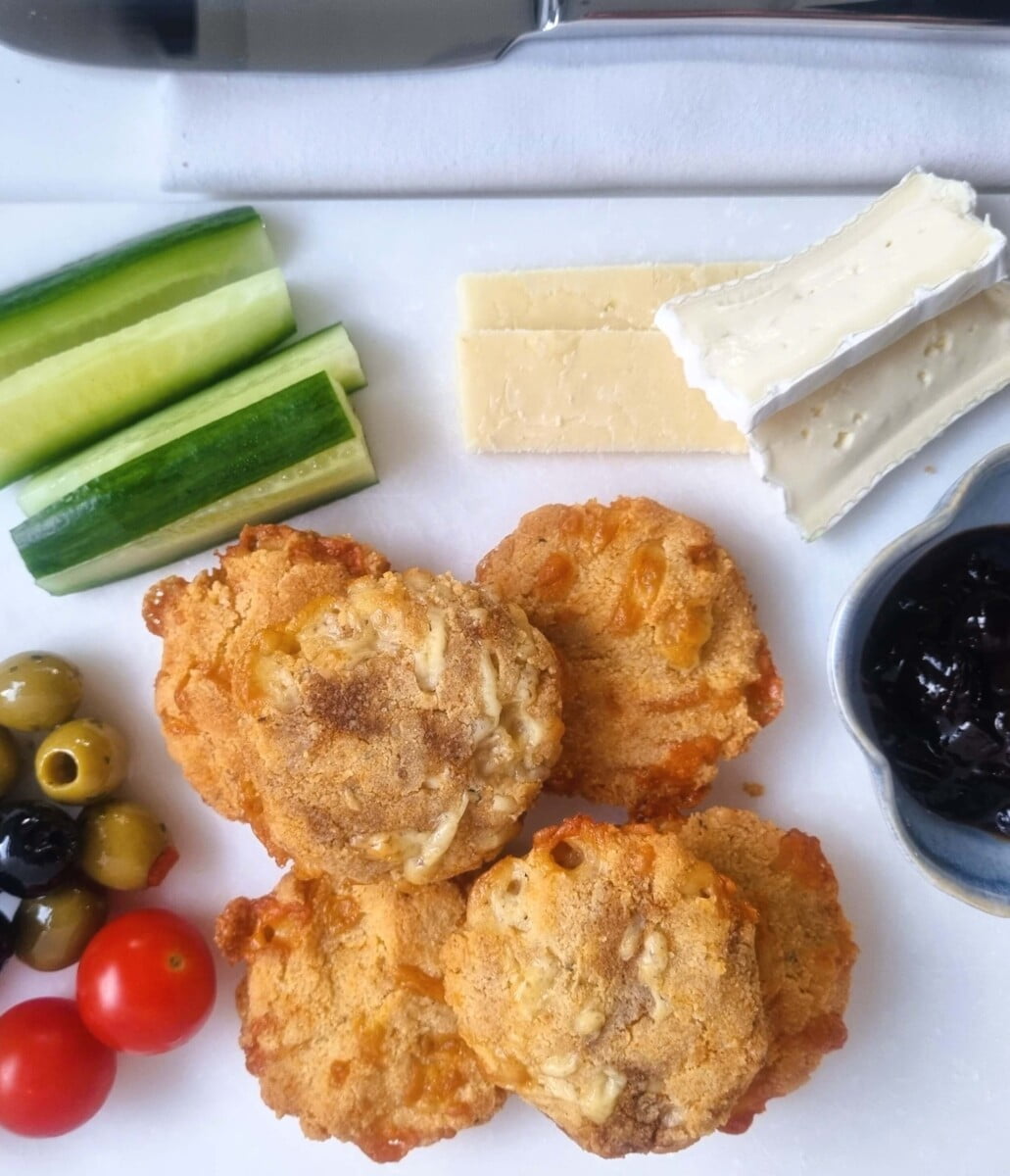 snack board of a stack of easy cheese  biscuits  with slices of cheese, chutney, cucumber, olives and tomatoes