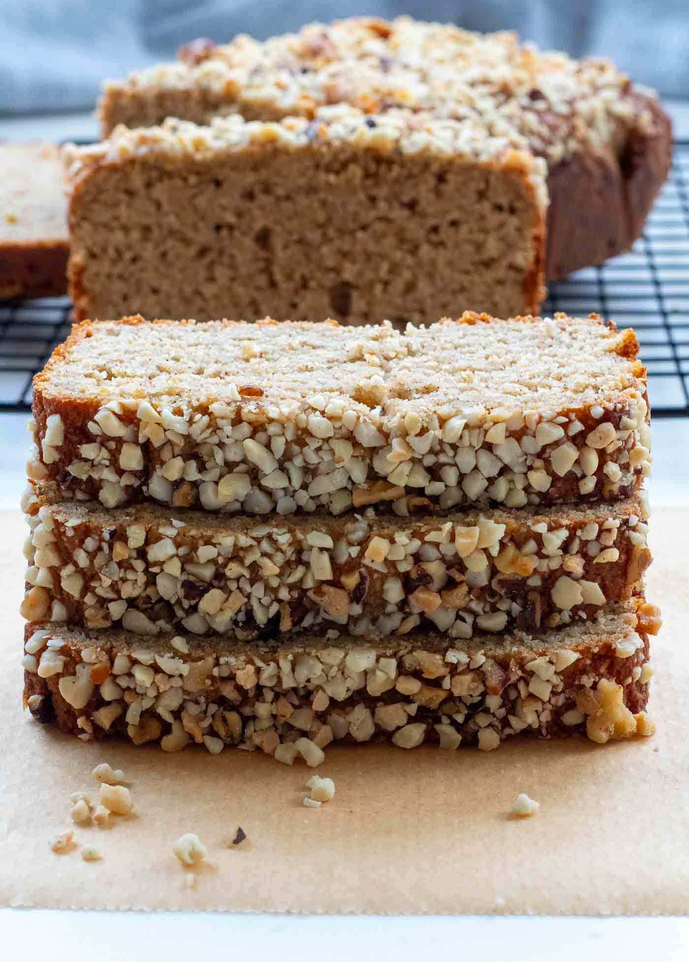 Slices of moist Healthy Diabetes-Friendly Banana Bread stacked on parchment paper, with chopped walnuts coating the edges. The bread has a soft, golden-brown crumb texture with a loaf in the background cooling on a wire rack.