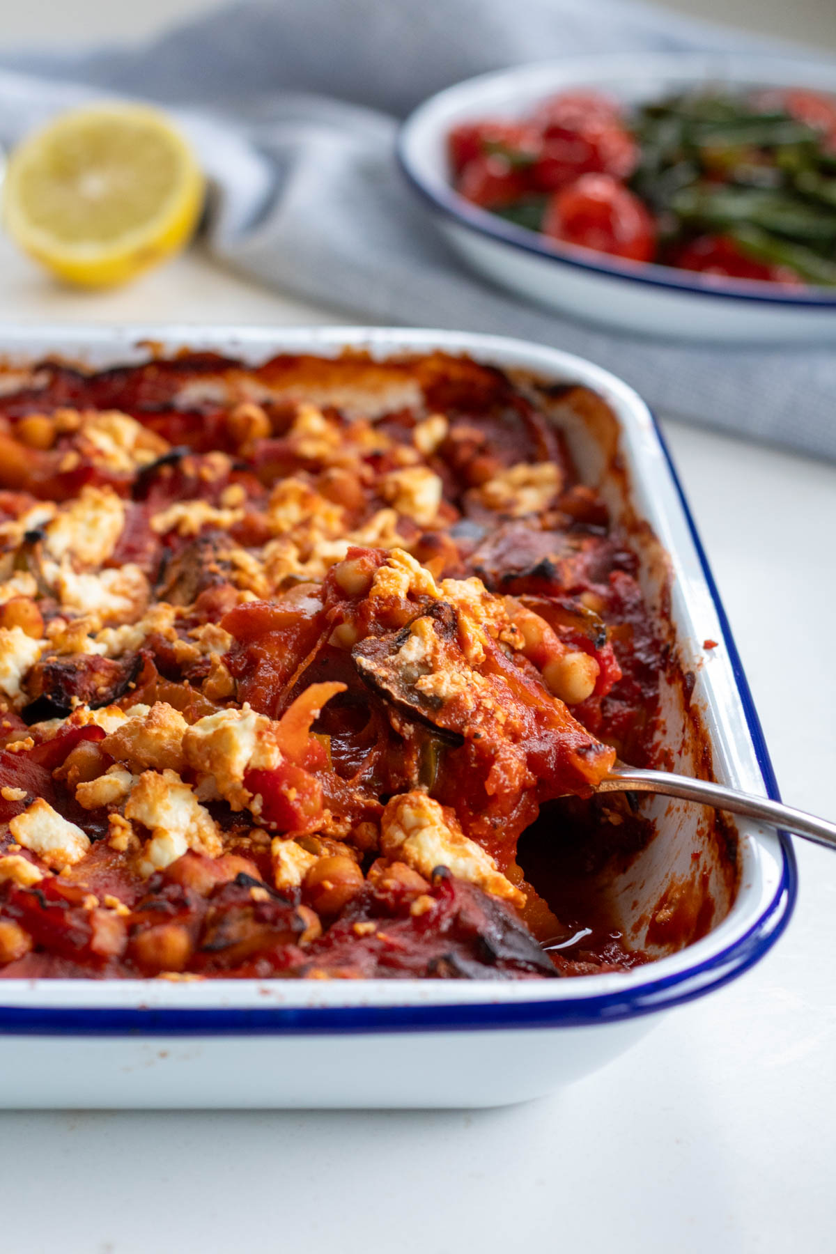 Aubergine Feta and Tomato bake in a roasting pan with a spoon lifting some out from the corner
