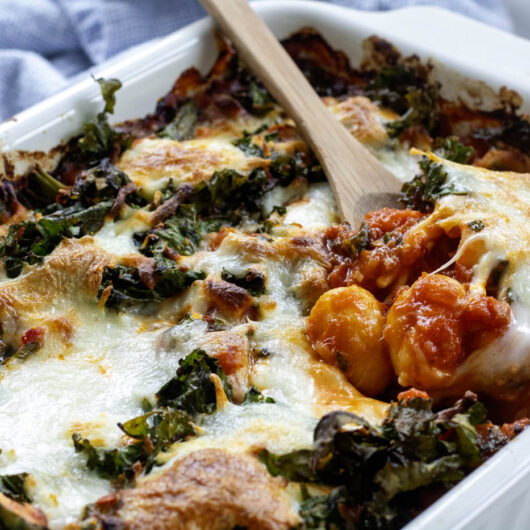 cooked vegetarian gnocchi bake with melted and crispy cheesey topping ina ceramic baking dish with a wooden serving spoon lifting some tomatoey gnocchi.