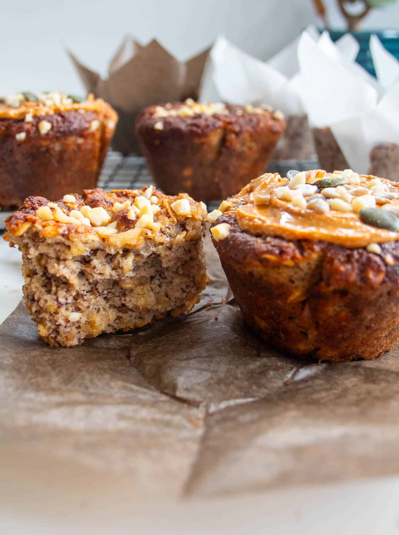 a closeup of a diabetes friendly banana oat muffin with a nut butter topping and chopped nuts and seeds sprinkled on top with anotehr muffin at the side cut in half on a brown muffin case wrapper. There are more muffins in the background in muffin cases