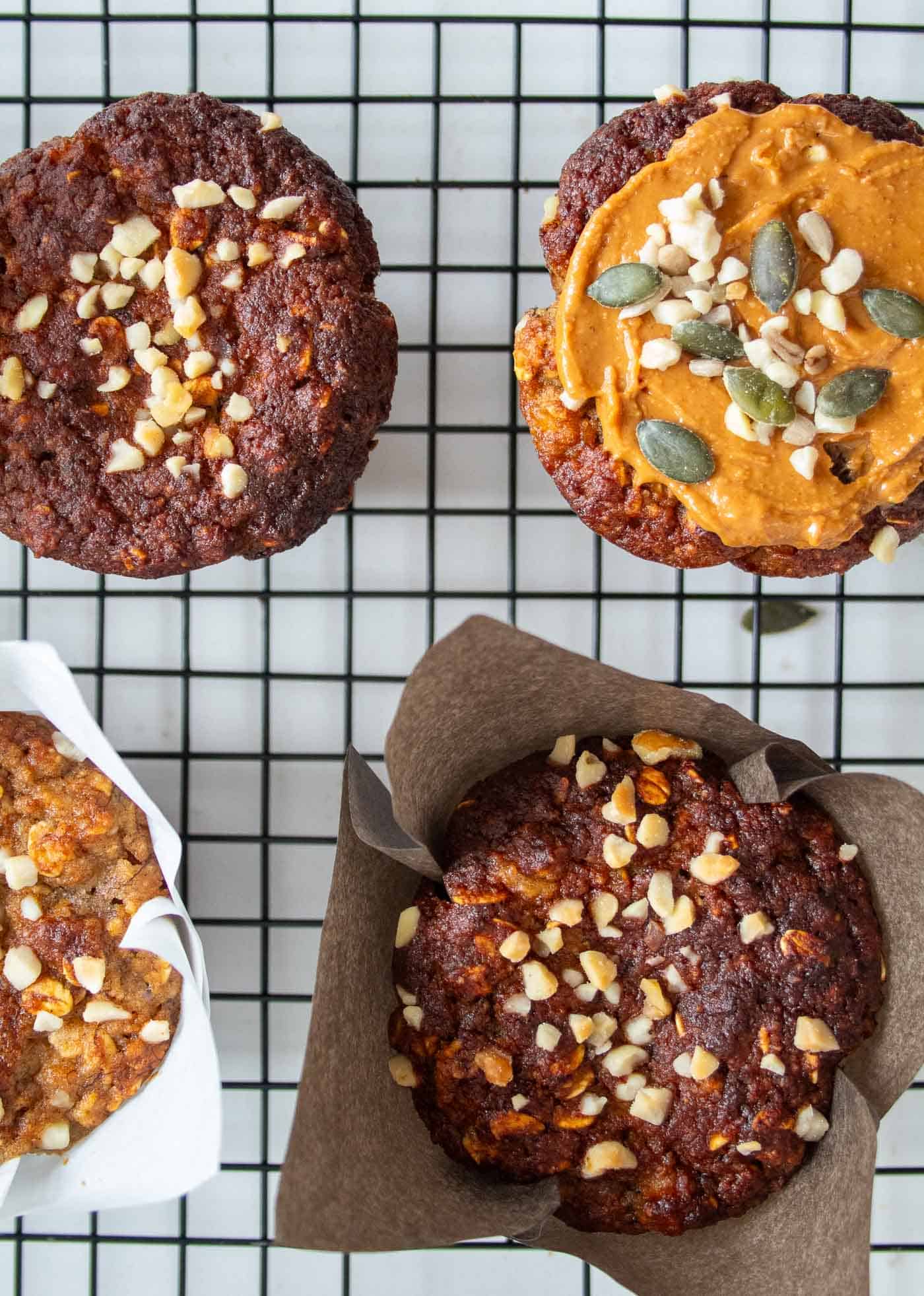 looking down on 4 healthy banana oat muffins on a black cooling tray with chopped nuts sprinkled on top. One banana and oat muffin has a topping of nut butter and seeds