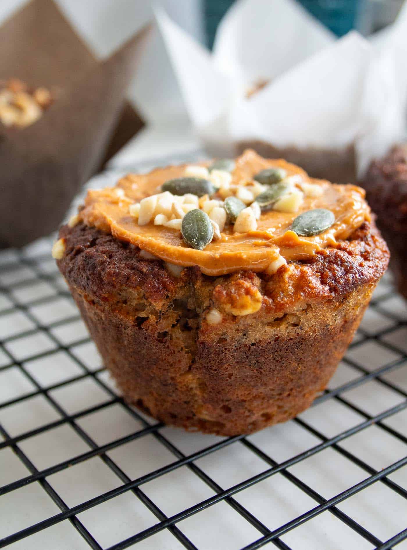 a closeup of a low sugar banana oat muffin on a cooling tray with a nut butter topping and chopped nuts and seeds sprinkled on top. There are more muffins in the background in muffin cases