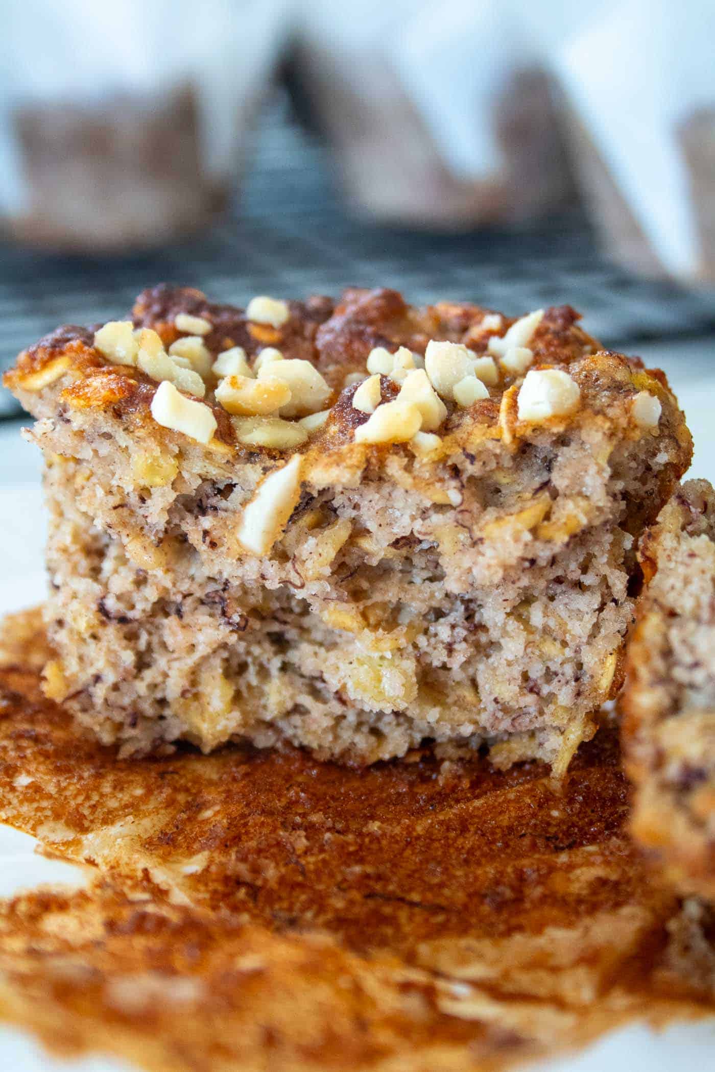 A closeup of half of a banana and oat muffin no sugar on a white muffin wrapper with chopped nuts on top. there are more muffins in the background on a black cooling tray