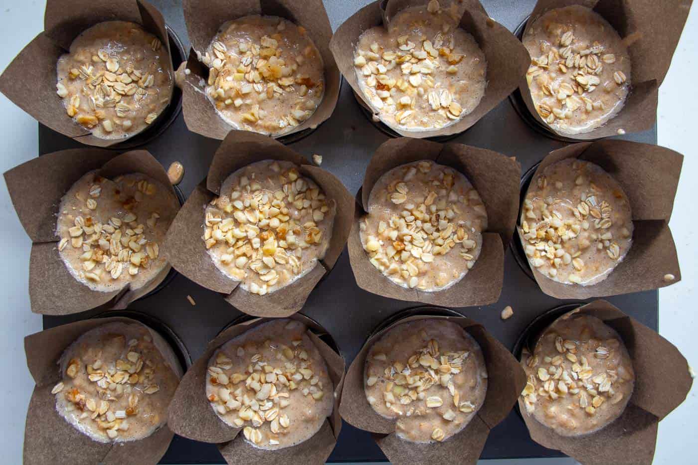 a muffin tin of uncooked banana and oat muffins in brown muffin cases with chopped nuts on top