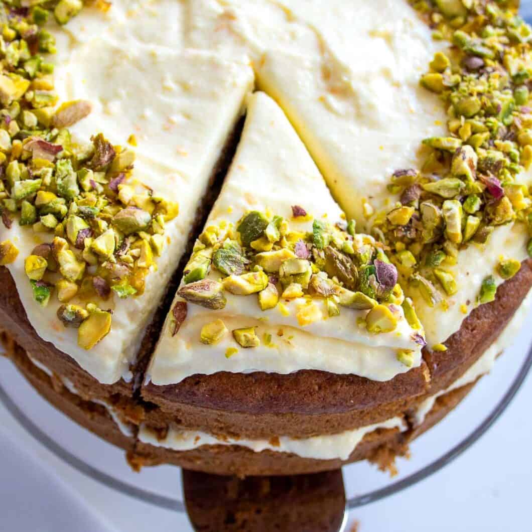 looking down on a sugar free carrot cake that has a cream cheese frosting on top with crushed pistachios around the edge. A slice has been cu and a silver cake slice is restin underneath