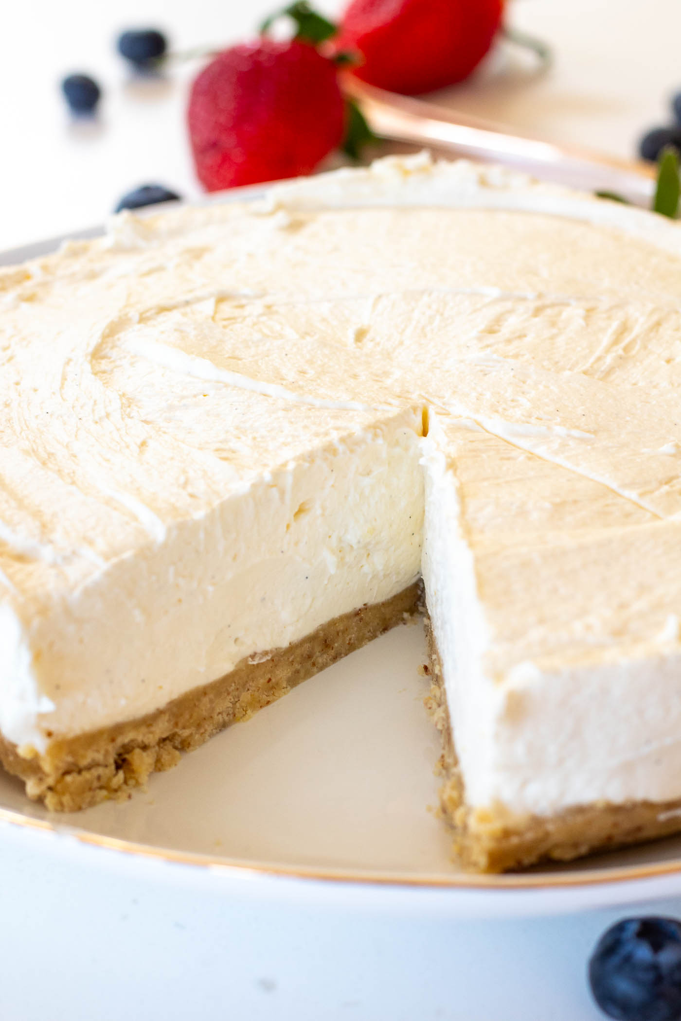 No-bake sugar-free vanilla mascarpone cheesecake with a slice missing, revealing its rich and creamy filling on a white plate witha gold rim and fresh blueberries and strawberries in the background