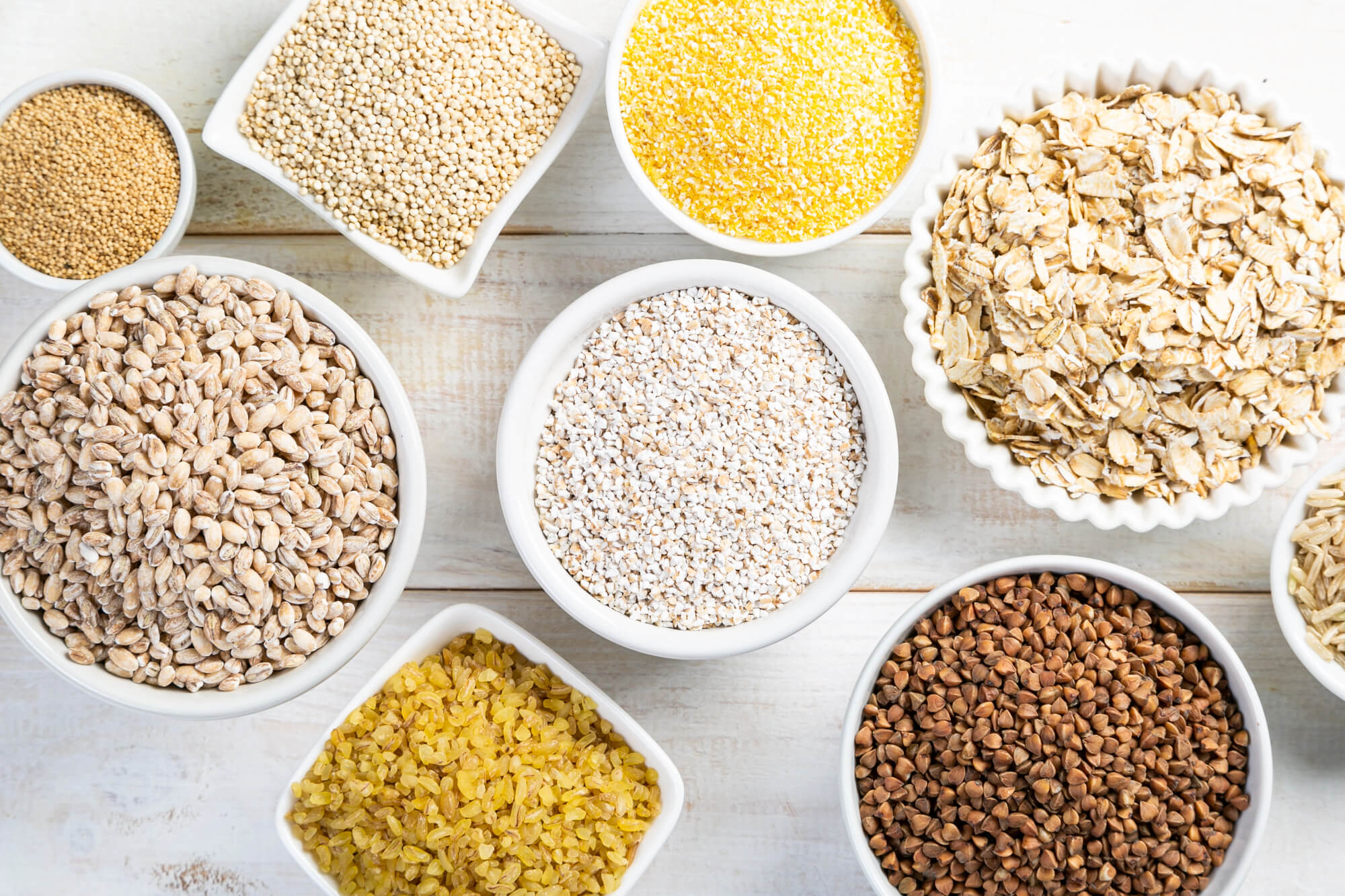 Selection of whole grains in white bowls - rice, oats, buckwheat, bulgur, porridge, barley, quinoa, amaranth.