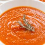 Smooth roasted butternut squash and tomato soup served in a white bowl with a sprig of rosemary on top.